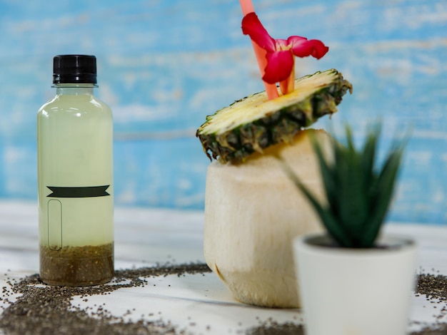 Close up shot of summer tropical coconut juice decorated with sliced sweet and sour pineapple blossom flower and straw placed on white and blue wooden table with bottle packaging for advertisement.