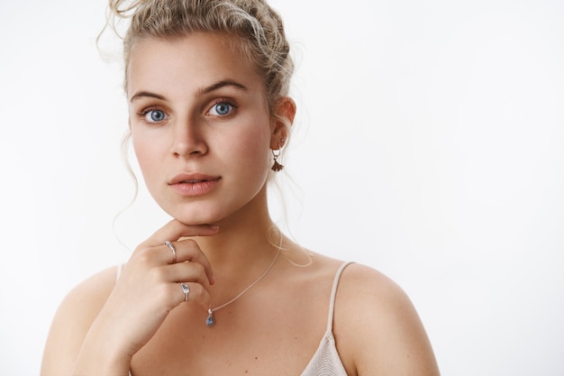 Close-up shot of thoughtful and sensual blond female european model with blue eyes in light dress touching chin soft and tender expressing tenderness and gentle feelings over white wall