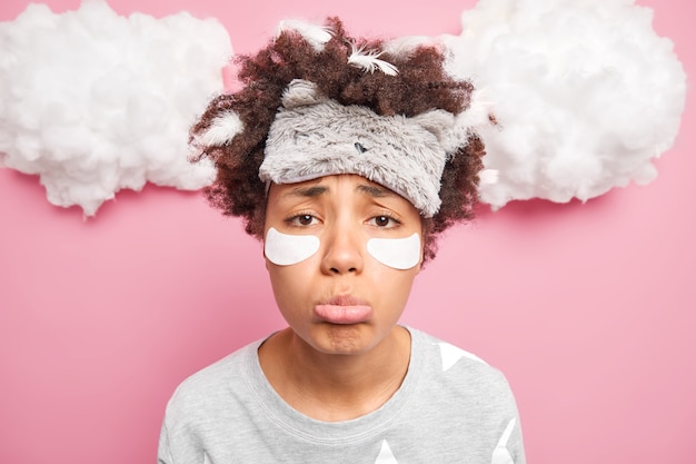 Photo close up shot of upset curly haired woman has frustrated unhappy look purses lower lip applies patches under eyes dressed in pajama and sleepmask isolated over pink wall