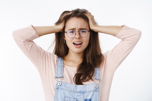 Close-up shot of upset uneasy and troubled woman in despair facing troublesome situation holding hands on head grimacing exhausted and displeased as panicking, posing concerned over gray wall.