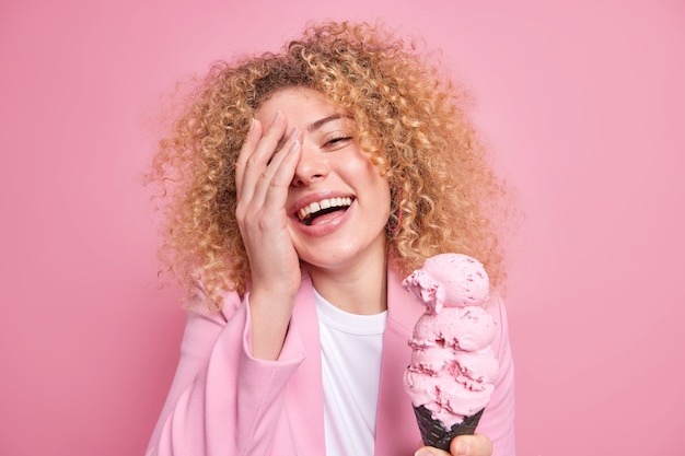 Close-up shot van dolblij krullende vrouw glimlacht breed maakt gezicht palm voelt geamuseerd houdt lekker smakelijk ijs in zwarte wafel geïsoleerd over roze muur. Mensen zomertijd junkfood