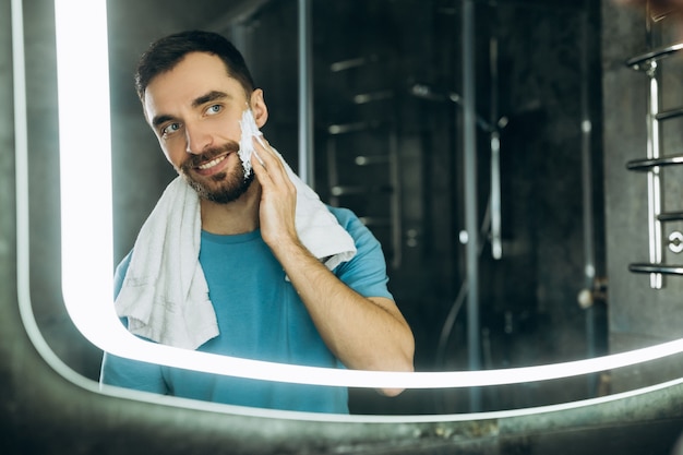 Close-up shot van een aantrekkelijke jonge man met een handdoek in zijn nek die voor de spiegel staat en 's ochtends vochtinbrengende crème op zijn gezicht in de badkamer aanbrengt.