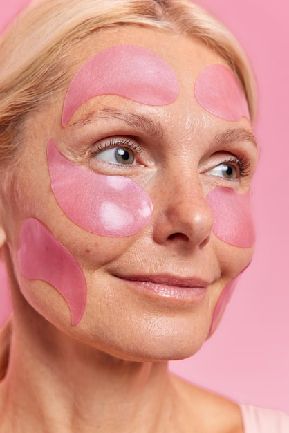 Close-up shot van een bedachtzame vrouw van middelbare leeftijd brengt roze vochtinbrengende pleisters op het gezicht aan om fijne lijntjes en rimpels te verwijderen, geconcentreerd in dagdromen op afstand over iets. Verjongingsconcept
