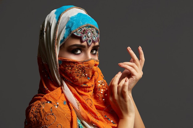 Close-up shot van een mooi meisje met professionele make-up, gekleed in een elegante kleurrijke hijab versierd met pailletten en sieraden. Ze gebaart en poseert zijwaarts in de studio en kijkt naar de