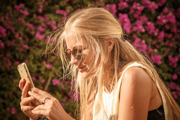 Close-up shot van een mooie jonge vrouw teksten op haar mobiele telefoon. Ze draagt een zonnebril en vrijetijdskleding. bloemen op achtergrond