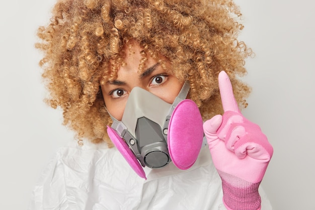 Close-up shot van een vrouw met krullend haar draagt een gasmasker en een beschermend pak steekt de vinger op heeft een idee hoe de verspreiding van het coronavirus te voorkomen, waarschuwt u voor gevaar geïsoleerd over grijze achtergrond besmetting