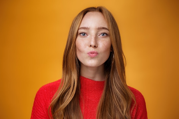 Close-up shot van flirterige knappe sensuele roodharige vrouw in rode gebreide kleding vouwen lippen in kus en staren naar de camera als het geven van mwah romantische en mooie gevoel tegen oranje achtergrond