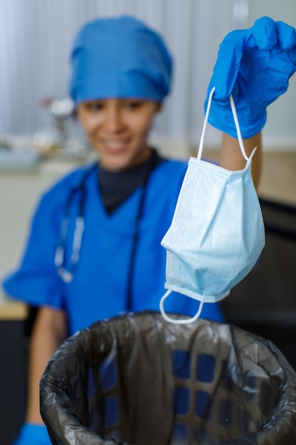 Close-up shot van gebruikt gezichtsmasker werd gedumpt in vuilniszak vuilnisbak door vrouwelijke gelukkig lachende arts in blauwe ziekenhuis uniforme rubberen handschoenen en stethoscoop op onscherpe achtergrond nadat pandemie eindigde.