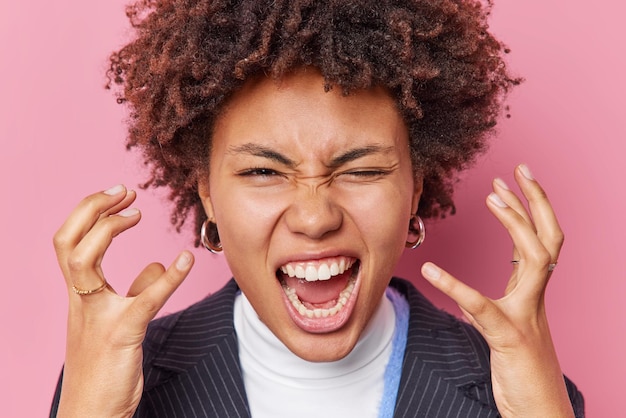 Close-up shot van geïrriteerde vrouw met krullend haar schreeuwt en gebaren boos geïrriteerd schreeuwt woedend voelt woedend en agressieve poses binnen tegen roze achtergrond negatieve emoties