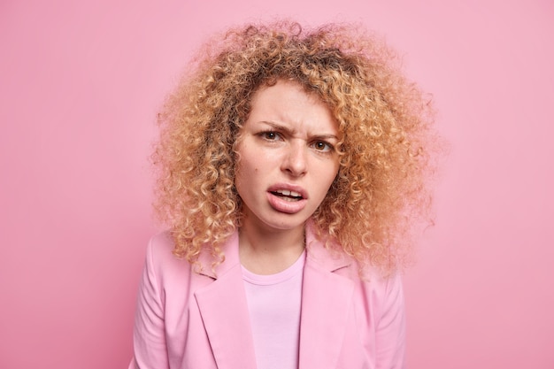 Close-up shot van gekrulde jonge vrouw ziet er ontevreden heeft verontwaardigde uitdrukking hoort onaangename woorden gericht ongelukkig gekleed in formele kleding geïsoleerd over roze muur