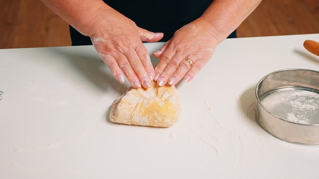 Close-up shot van handen van senior bakkerij deeg kneden. Gepensioneerde oudere bakker met bonete die ingrediënten mengt met gezeefd tarwemeel dat zich vormt op een met bloem bestoven oppervlak voor het bakken van traditionele cake, brood