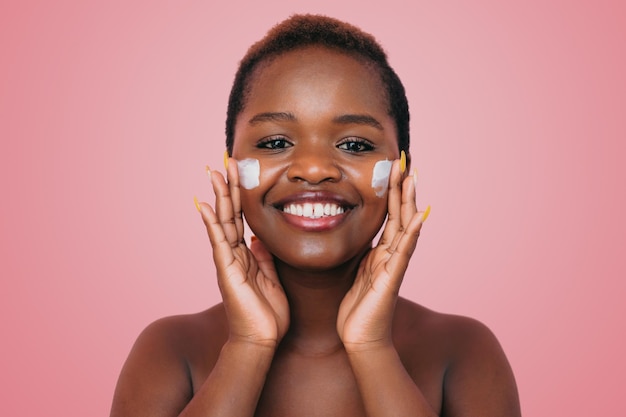 Foto close-up shot van positieve donkere vrouw die hydraterende crème op haar gezicht aanbrengt geïsoleerd op roze achtergrond huidverzorging en beauty concept dermatologie cosmetologie