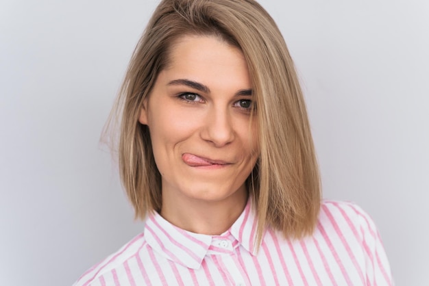 Close-up shot van positieve grappige blonde vrouw met aantrekkelijke look en mooie groene ogen dragen roze shirt met tong poseren tegen witte muur in studio Gelukkige vrouw met positieve emoties