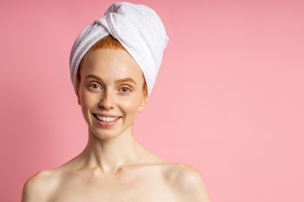 Close-up shot van prachtige gelukkige blanke roodharige vrouw met schone, gezonde huid, staande met witte handdoek op het hoofd na het douchen op roze achtergrond. Lichaamsverzorgingsconcept, kopieer ruimte
