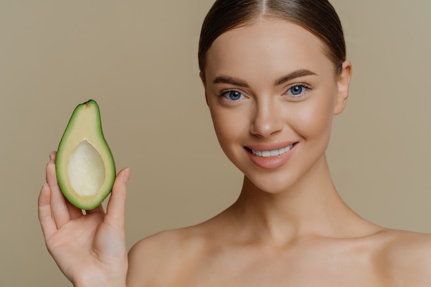 Close-up shot van topless jonge blanke vrouwelijke model heeft donker gekamd haar glimlacht zachtjes houdt de helft van avocado stands shirtless indoor geeft om huid en teint. Schoonheid wellness-concept