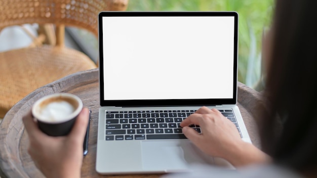 Photo close-up shot of woman using a blank screen laptop and holding a coffee mug shot from the back