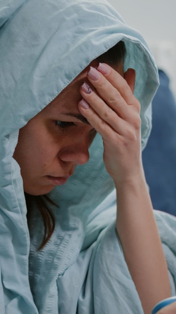 Close up of sick woman holding thermometer and measuring temperature while sitting in blanket. Caucasian adult feeling cold and shivering, checking fever diagnosis on medical device
