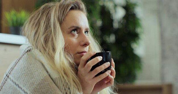 Close up of sick woman sitting at home in blanket with hot tea\
suffering from fever temperature infection flu headache cold. young\
person drinking tea, with seasonal virus symptoms feeling\
unwell.