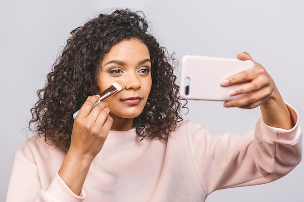 Close up side portrait of beautiful aro american woman applying makeup with brush. Foundation for dark skin. Using phone.