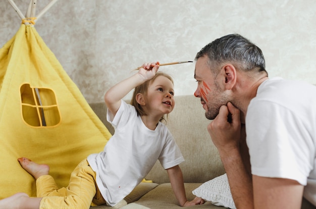 Foto vista laterale del primo piano di una bambina felice che applica il pennello sul viso di suo padre. un bel padre maschio sorridente gioca con sua figlia sul divano di casa.