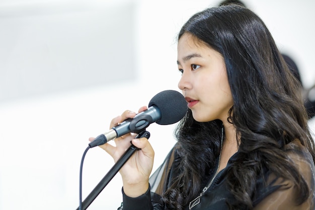 Close-up side view portrait shot of asian teenage singer\
singing a song with a microphone. happy junior student vocalist\
practicing in studio. beautiful girl rehearsing to prepare for the\
competition