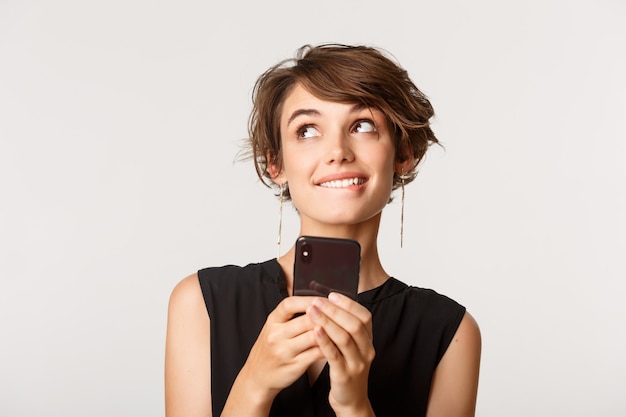 Close-up of silly girl looking upper left corner dreamy, standing with smartphone over white.