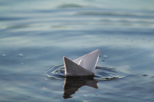 Primo piano di piccola barca bianca semplice della carta di origami che galleggia tranquillamente in chiaro fiume blu o acqua di mare sotto il cielo luminoso di estate. concetto di libertà, sogni e fantasie, sfondo copyspace.