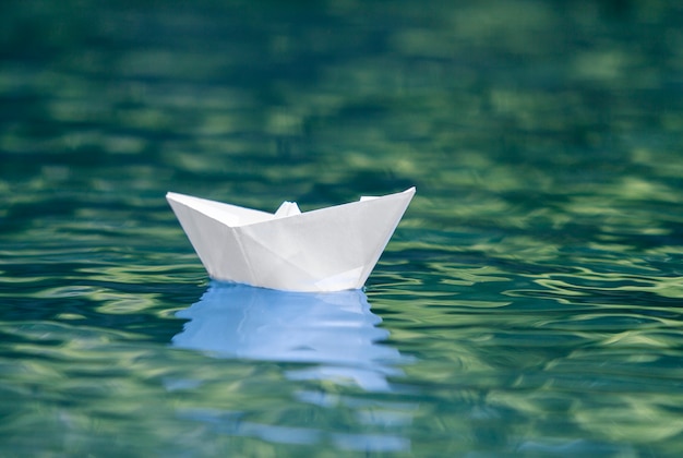 Close-up of simple small white origami paper boat floating quietly in blue clear river or sea water under bright summer sky. 