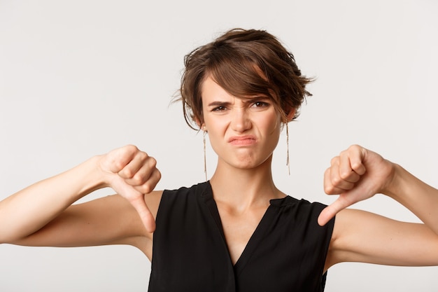 Close-up of skeptical and disappointed woman grimacing, showing thumbs-down, express dislike, negative review, standing over white.