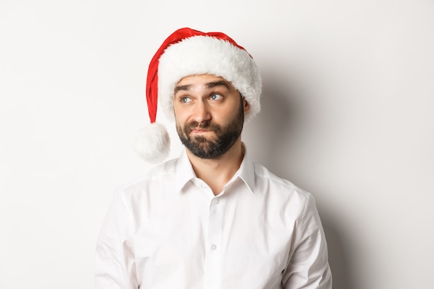 Close-up of skeptical guy in santa hat looking doubtful left, grimacing with hesitation, white background.