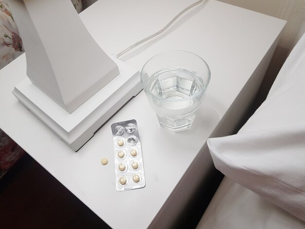 健康と病気の概念である寝室のベッドサイドテーブルでの穏やかで健全な睡眠のために準備された睡眠薬とコップ一杯の水のクローズアップ。