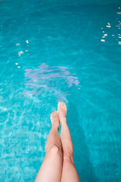 Close-up of slender female legs against the blue water by the pool on a hot summer day, under the sun. The girl dangles her legs in the water of the pool, resting, sunbathing.