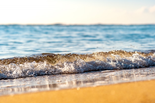 Foto chiuda su di piccole onde del mare con chiara acqua blu sopra la spiaggia di sabbia gialla alla riva soleggiata dell'estate.