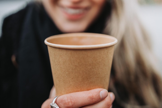 曇りの日の午後に使い捨てのコーヒー カップを保持している笑顔の金髪女性のクローズ アップ。モックアップ