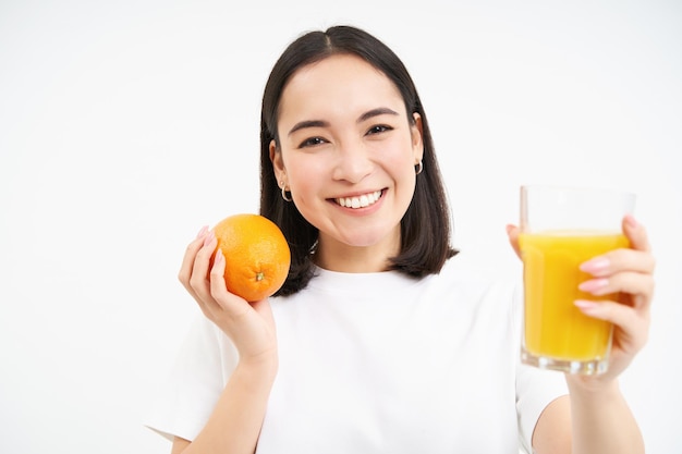 Close up of smiling brunette girl drinks fresh juice holds orange fruit white studio background