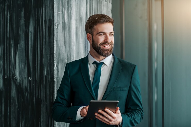Photo close up of smiling caucasian businessman in formal wear using tablet and looking away while standing next to wall.