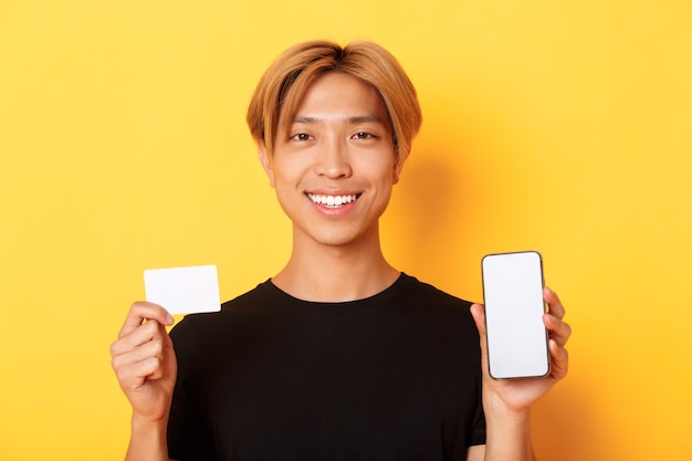 Close-up of smiling handsome asian guy showing smartphone screen and credit card