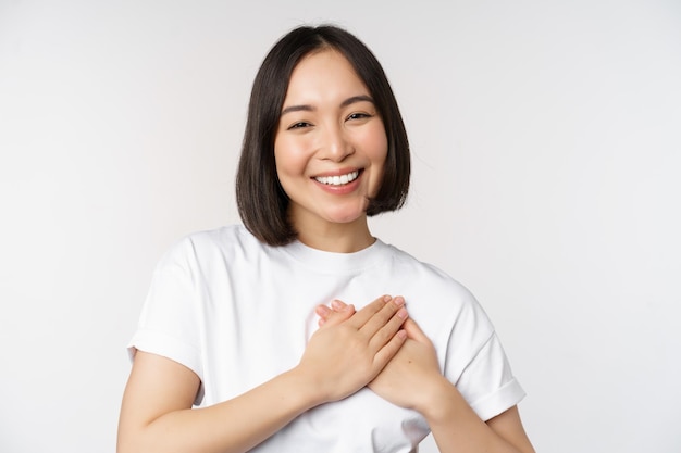 Close up of smiling korean woman holding hands on heart care and love concept feel affection tenderness or heartwarming feeling standing over white background