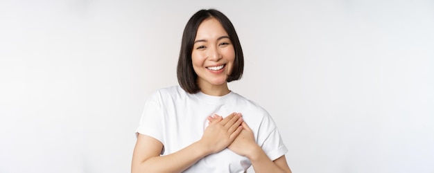 Close up of smiling korean woman holding hands on heart care and love concept feel affection tenderness or heartwarming feeling standing over white background