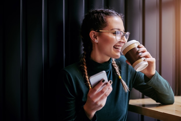 Photo close up of smiling teenage girl sitting in cafeteria next to window, drinking coffee and holding smart phone while looking trough window.