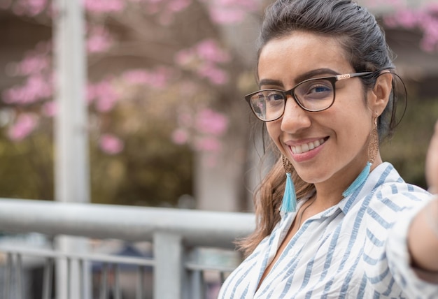 Close up of smiling woman taking a selfie with a beautiful tree in background at urban scene