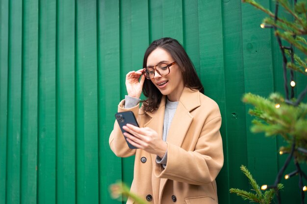 Close up smiling woman using smartphone standing near christmas tree