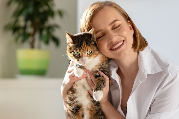 Close up of smiling woman in white shirt hugging and embracing with tenderness and love domestic cat in home