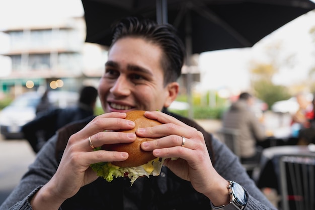 Photo close-up smiling young man holding a gourmet burger sitting at table.