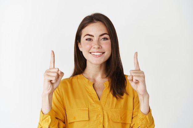 Photo close-up of smiling young woman showing logo, pointing fingers up and looking confident at camera, standing over white wall