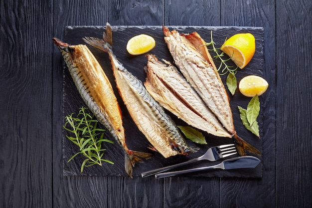 Close-up of smoked mackerel served on a black slate tray with salt, peppercorn, rosemary and lemon slices, fork and knife, view from above