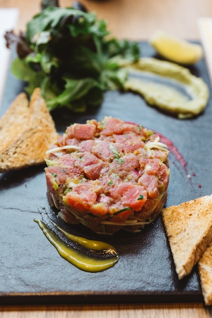 Close up tartare di tonno rosso piccante con salsa agrodolce e piccante. servito con pane tostato e insalata su lastra di pietra nera.