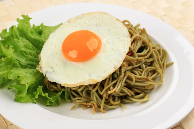 Close Up Spicy Healthy Green Instant Fried Noodle with Shiny Sunny Side Egg and Lettuce on White Ceramic Round Plate.