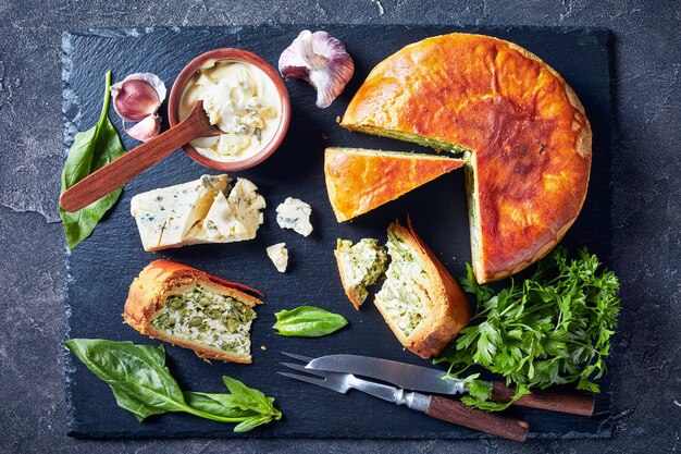 Close-up of spinach Green Bean and blue cheese pie sliced on a black slate tray with blue cheese cream sauce in a bowl on a concrete table, horizontal view from above