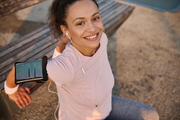 Close-up of sportswoman female athlete in pink t-shirt, with\
earphones and smartphone holder exercising outdoor, flexing her\
triceps on wooden bench, smiling toothy smile confidently looking\
at camera